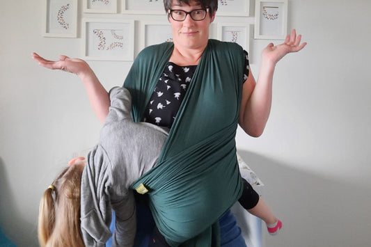 woman with her hands in the air, wearing a toddler who is leaning out of a green stretchy wrap