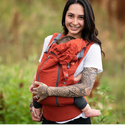 Women with sleeping baby in rust coloured Beco8 carrier, the hood is up to support the childs head.