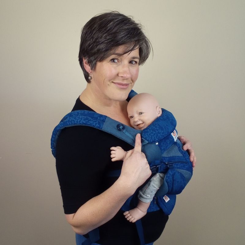 Joy Riders Babywearing Consultant Sarah wearing a demonstration doll in a Blue Ergobaby Omni 360 Carrier. Sarah is pointing at the baby and smilling