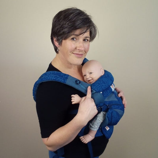 Joy Riders Babywearing Consultant Sarah wearing a demonstration doll in a Blue Ergobaby Omni 360 Carrier.  Sarah is pointing at the baby and smilling